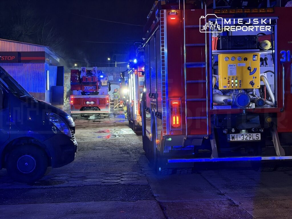 Czerwony wóz strażacki z włączonymi sygnalizatorami świetlnymi stoi na parkingu, obok niego strażacy przy pracy.