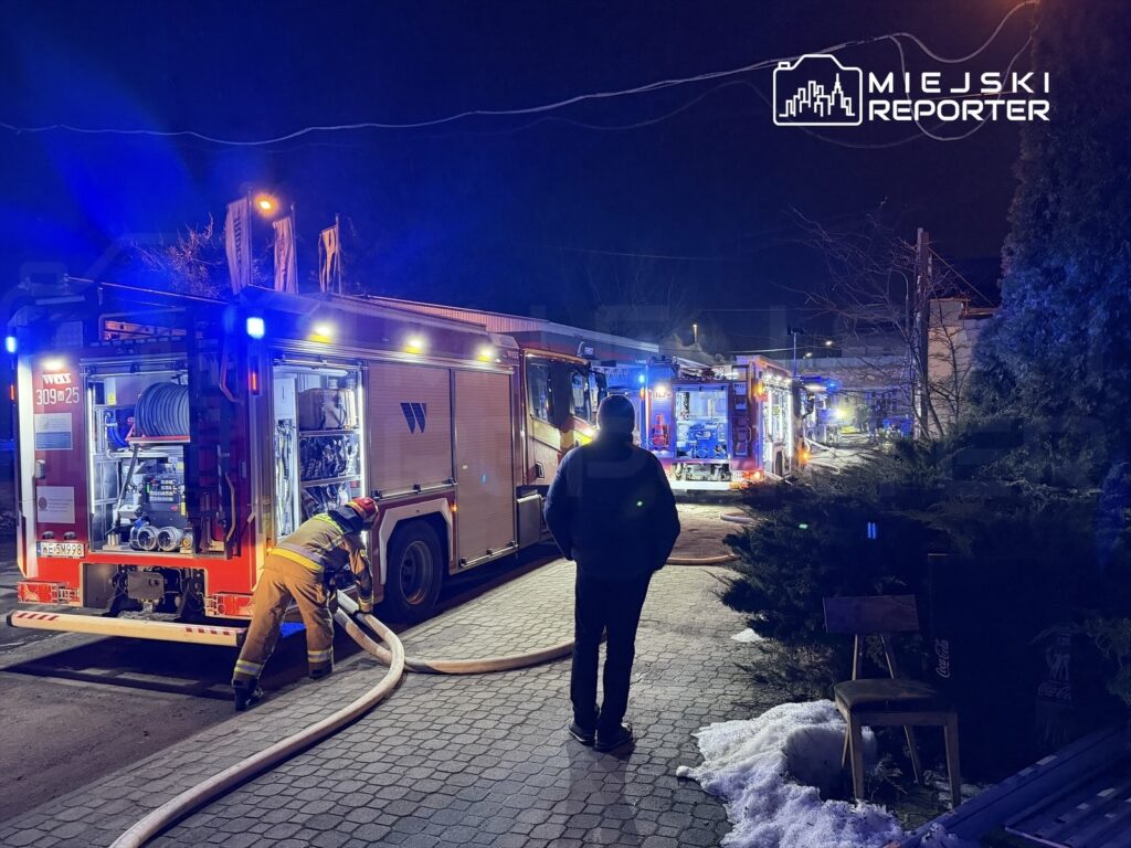 Strażak w mundurze podłącza wąż do wozu strażackiego na oświetlonej ulicy w nocy, obok stoją inne pojazdy ratunkowe.