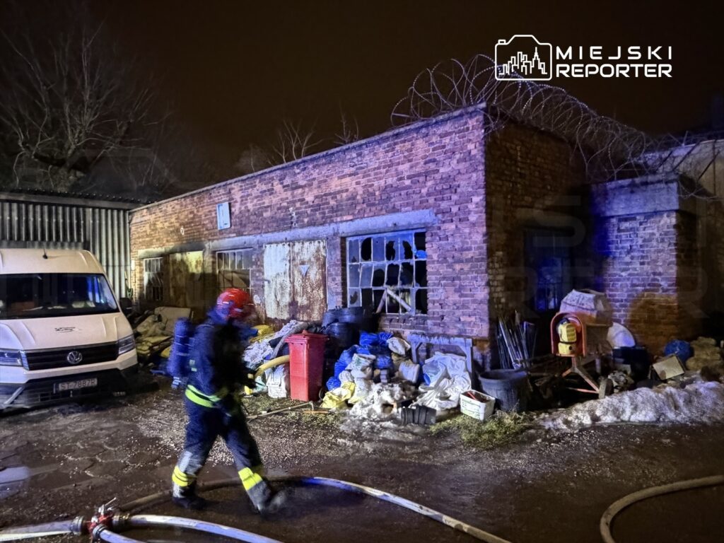 Strażak w hełmie i mundurze przechodzi obok spalonego budynku z uszkodzonymi oknami, w tle widoczne narzędzia i odpady.