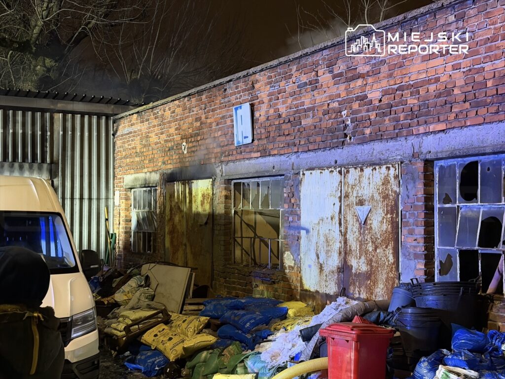 Zniszczony budynek z cegły, z widocznymi oknami i metalowymi drzwiami, otoczony odpadami i workami.