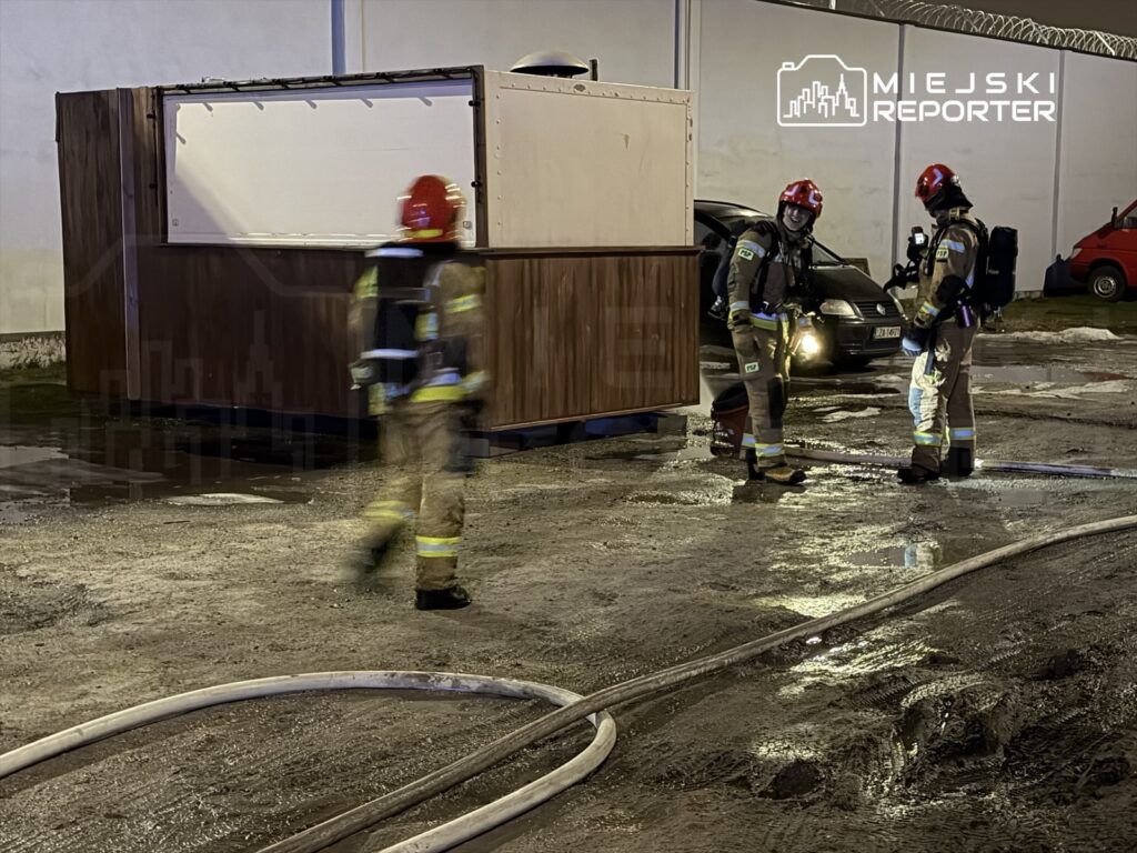 Strażacy w hełmach i mundurach omawiają sytuację obok drewnianej konstrukcji w mokrej okolicy po akcji ratunkowej.