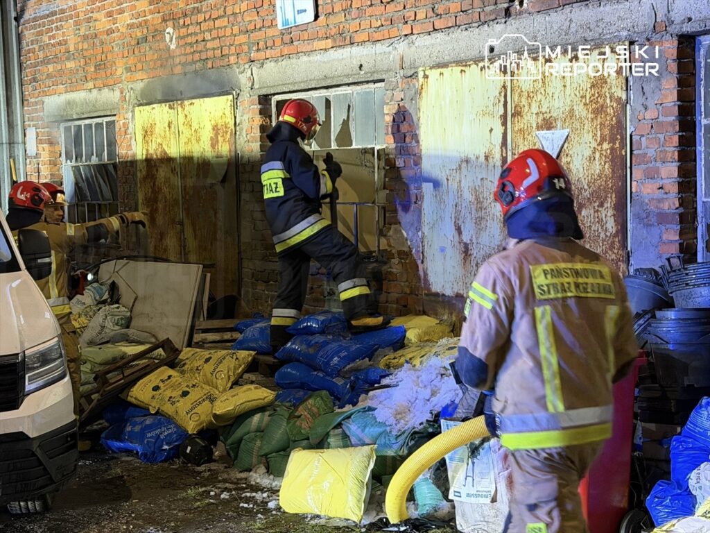 Dwóch strażaków w hełmach przeszukuje zniszczony budynek, otaczają ich worki z materiałami budowlanymi.