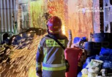 Strażak w kasku i mundurze obserwuje iskry wydobywające się z płonącego materiału w zniszczonym budynku.