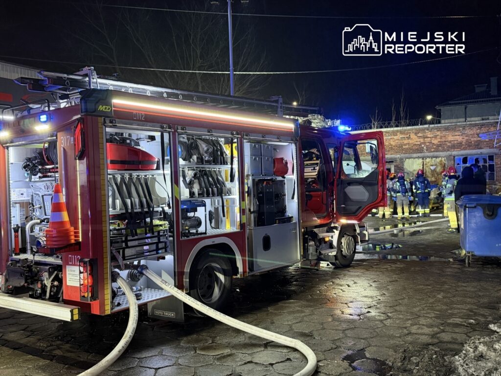 Wozidło strażackie z otwartą tylną częścią, z wężami pożarniczymi, w tle strażacy pracujący przy budynku.