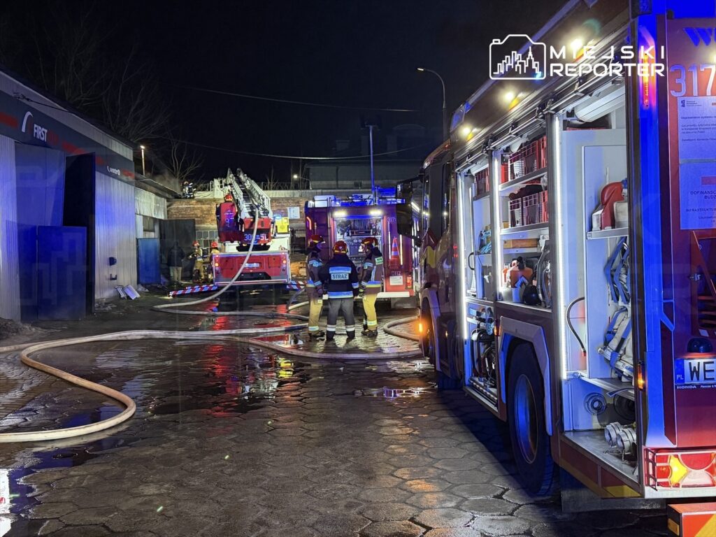 Strażacy w mundurach pracują przy wozach strażackich, podając węże gaśnicze w oświetlonym, nocnym otoczeniu.
