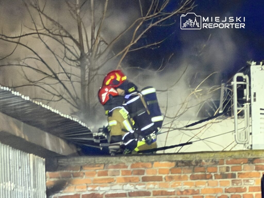 Strażak w hełmie gaszącym pożar na dachu budynku, w tle widoczne unoszące się kłęby dymu.