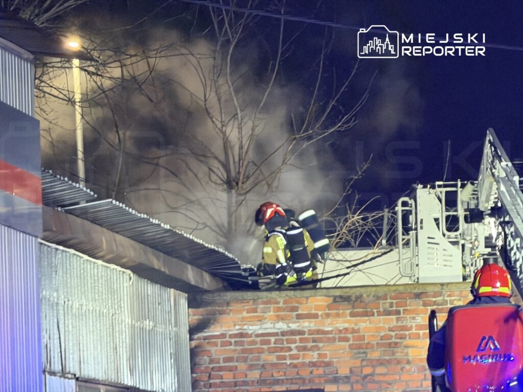Strażacy gaszą pożar na dachu budynku, w tle widoczne kłęby dymu i oświetlenie uliczne.