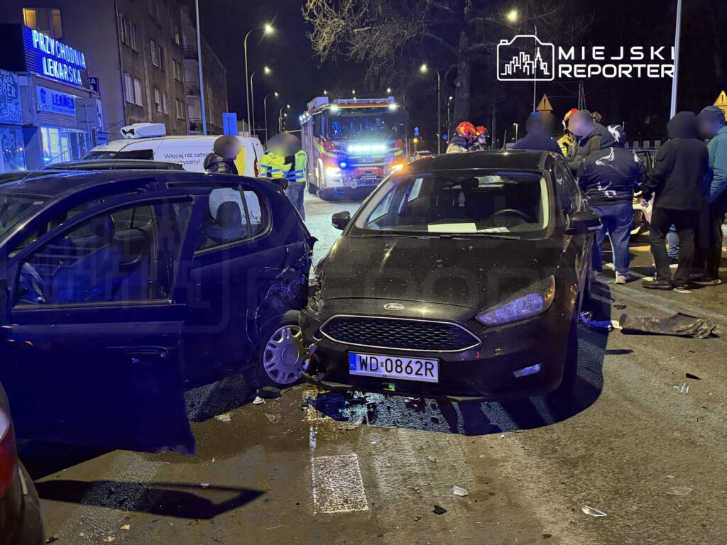 Dwa uszkodzone samochody na ulicy, z otwartymi drzwiami jednego z pojazdów, obok grupka ludzi oraz obecność służb ratunkowych.