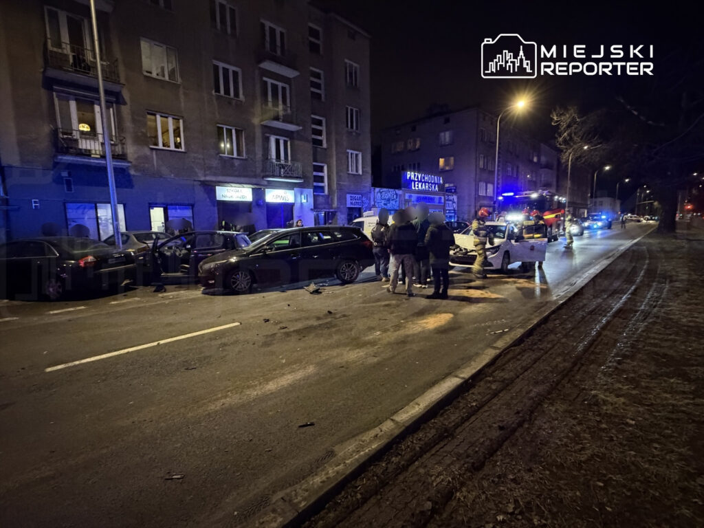 Na ulicy w nocy grupa ludzi zbiera się wokół zderzonych samochodów, wokół widać ślady zderzenia i interwencję służb.