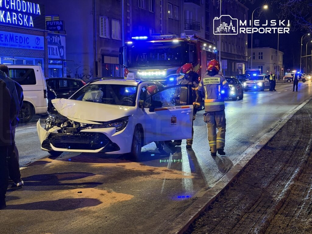 W nocy na ulicy doszło do wypadku, w którym uszkodzony biały samochód osobowy jest otoczony przez strażaków oraz policję.