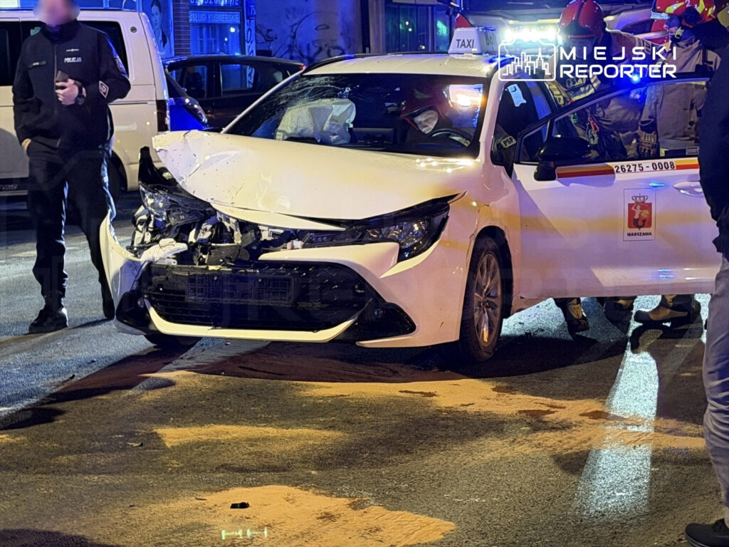 W nocy, w mieście, uszkodzony biały samochód osobowy stoi na jezdni, otoczony przez policjantów i strażaków.