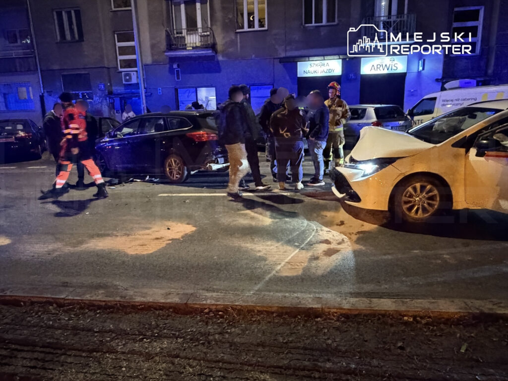 Na ulicy grupa osób w odzieży roboczej oraz mundurach rozmawia obok uszkodzonego samochodu. W tle widać policyjne światła.