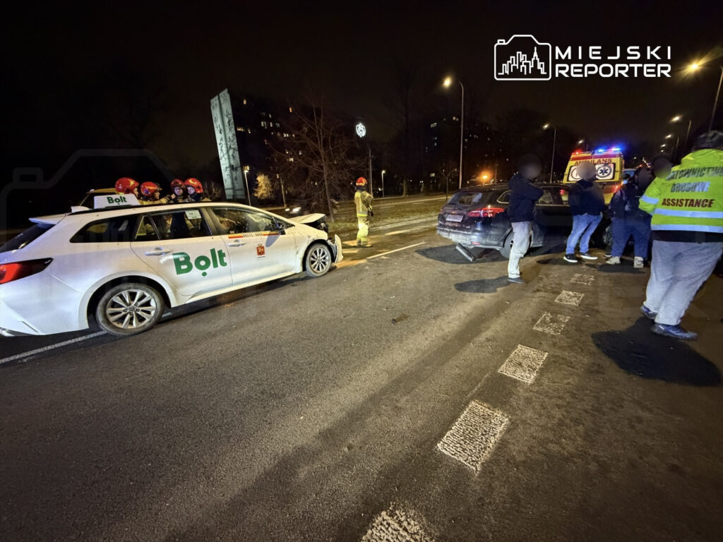 Na drodze stoi biały samochód osobowy z logo Bolt, obok grupka osób oraz zespół ratunkowy udzielający pomocy.