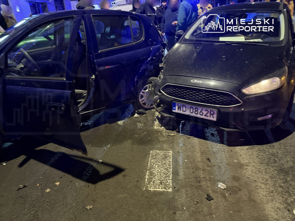 Dwa uszkodzone samochody osobowe po kolizji na ulicy, przy jednym z otwartych drzwi pojazdu. W tle zebrani ludzie.