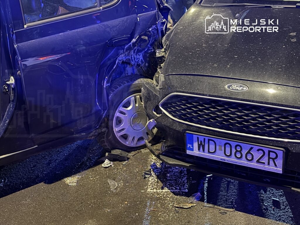 Dwa uszkodzone samochody osobowe, jeden czarny Ford z widocznym uszkodzeniem, stoją na ulicy w nocy.