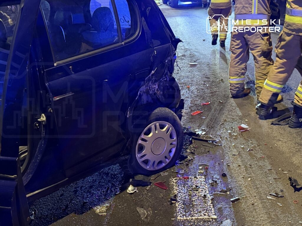 Uszkodzony samochód osobowy z otwartymi drzwiami stoi na ulicy, obok pracują strażacy.