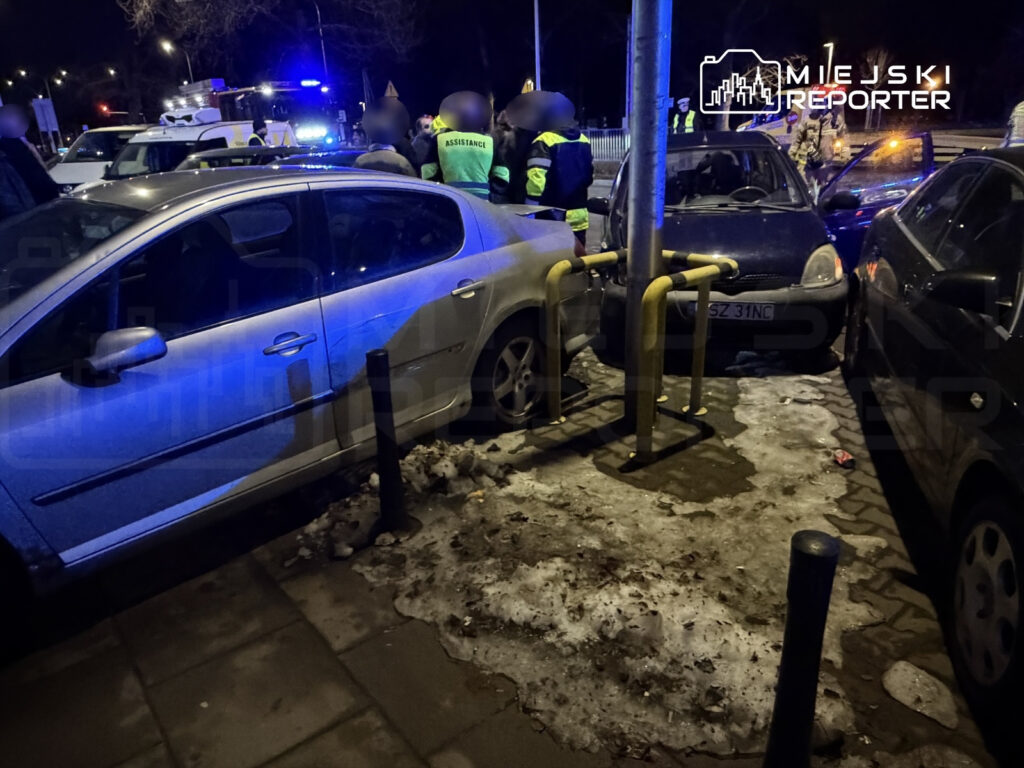 Grupa osób w odblaskowych kamizelkach interweniuje przy zderzeniu dwóch samochodów na oświetlonej ulicy.