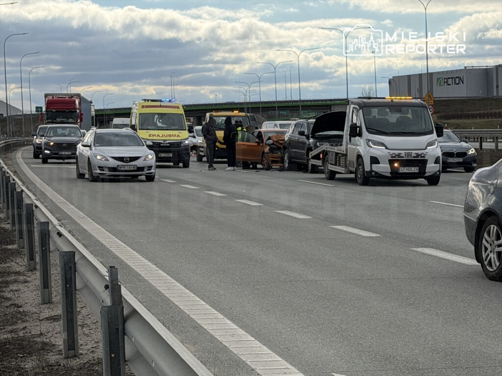 Na drodze stoi kilka pojazdów, w tym karetka i auto lawetowe, a obok nich grupka osób rozmawia przy uszkodzonym samochodzie.