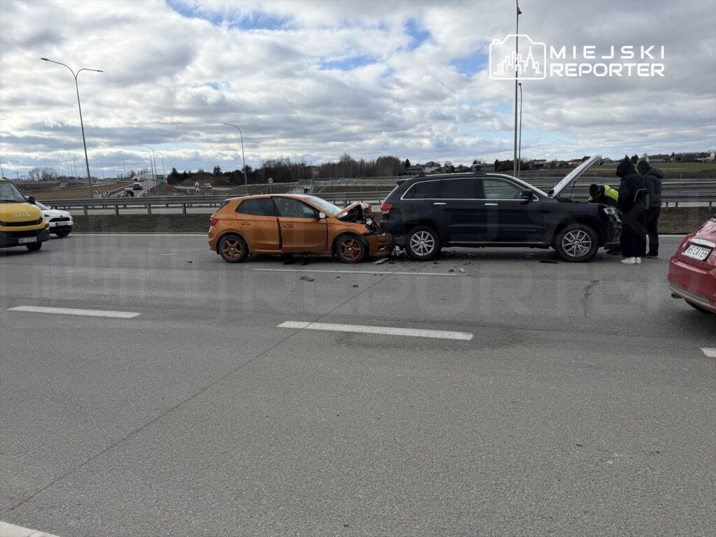 Dwa uszkodzone samochody, pomarańczowy SUV i czarny SUV, na drodze, z osobami sprawdzającymi stan pojazdów.