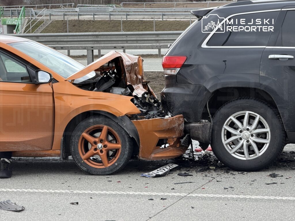 Zderzenie dwóch samochodów na drodze, z widocznym uszkodzeniem przedniej części pomarańczowego auta i tyłu czarnego SUV-a.
