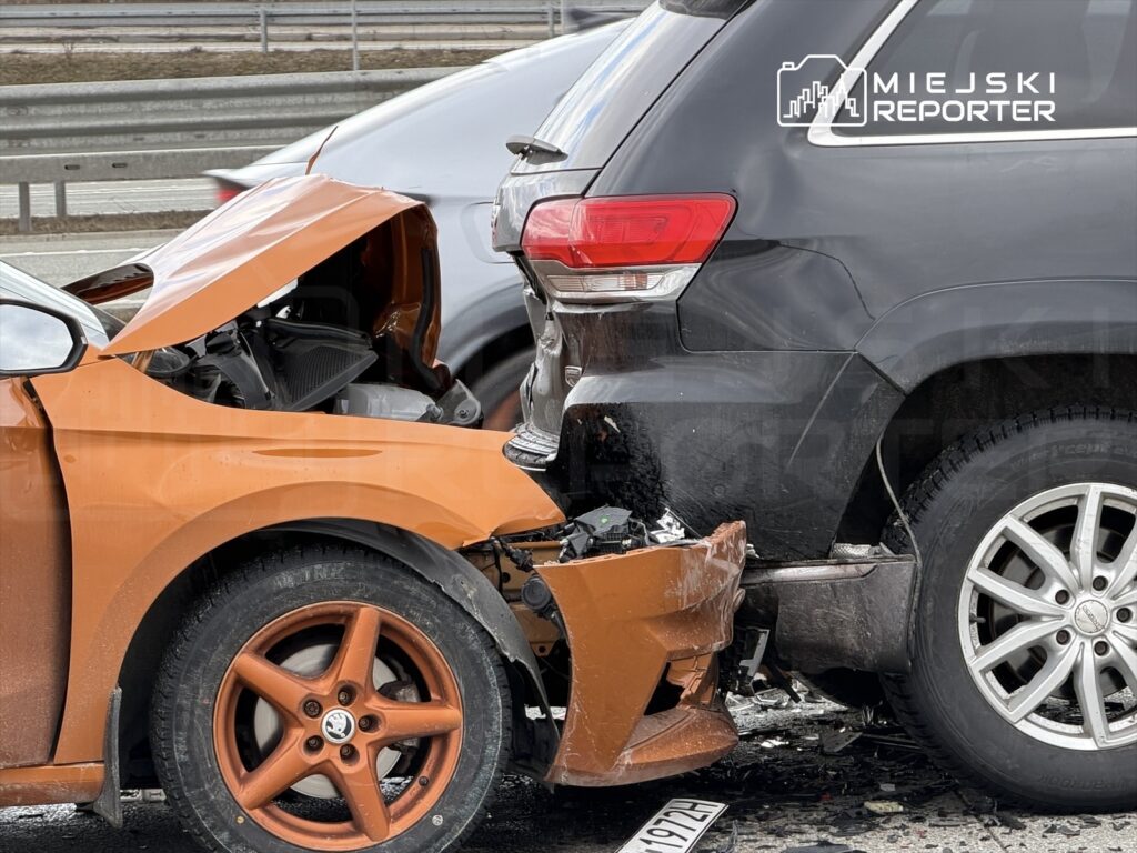 Zderzenie dwóch samochodów osobowych na drodze, z widocznymi uszkodzeniami tyłu czarnego SUV-a i przodu pomarańczowego pojazdu.