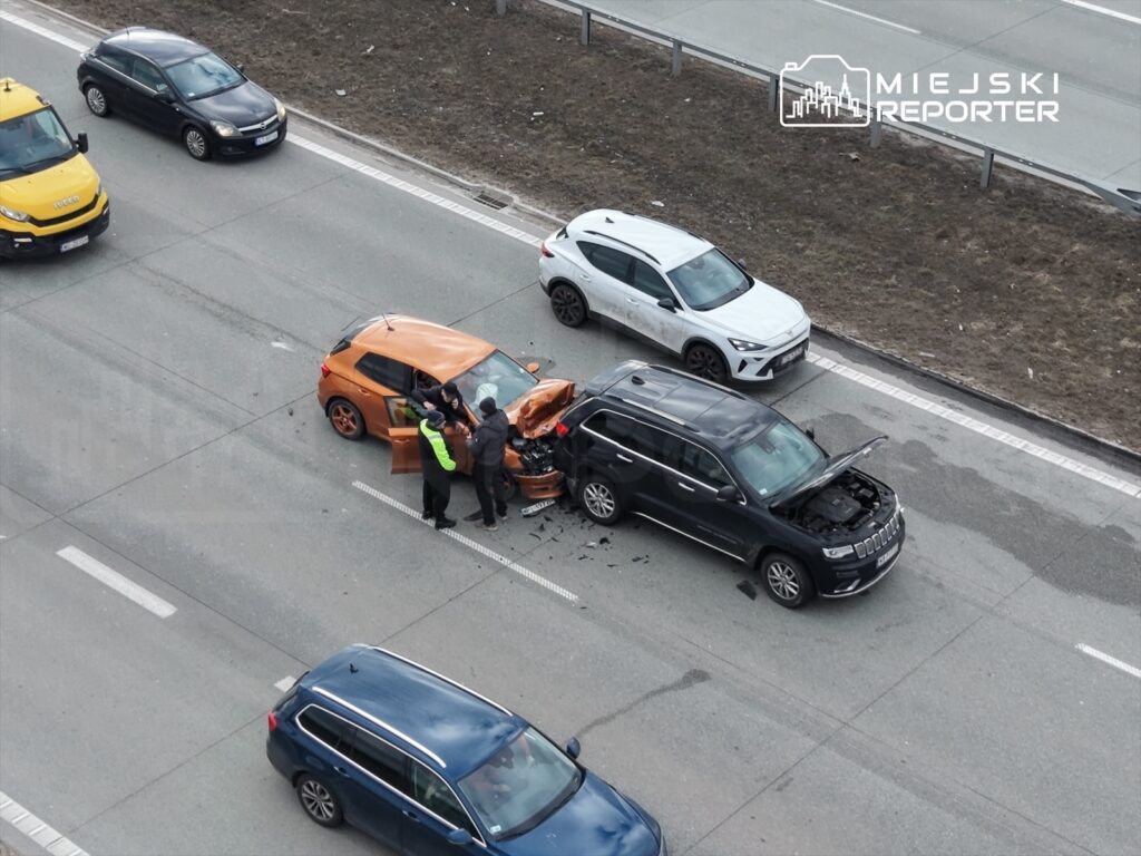 Wielu mężczyzn w odblaskowych kamizelkach bada uszkodzone samochody na drodze, obok przejeżdżające pojazdy.