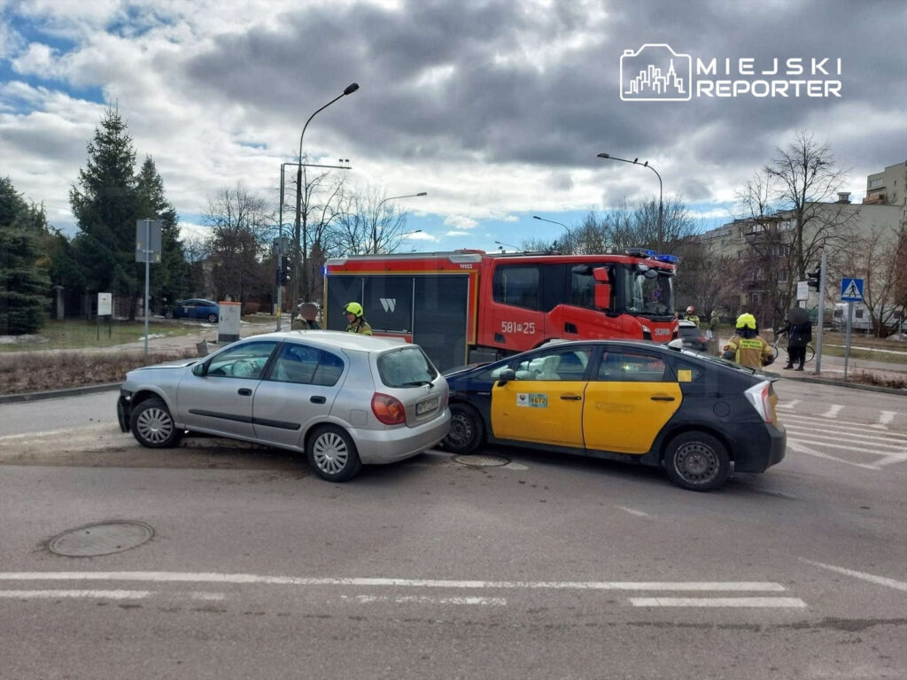 Dwa uszkodzone samochody, jeden srebrny, drugi czarny z żółtym oznaczeniem, zderzyły się na skrzyżowaniu; obok strażacy w akcji.