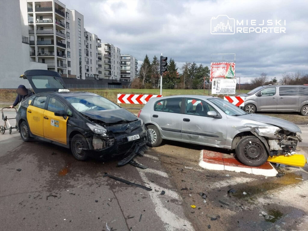 Dwa uszkodzone samochody osobowe, taksówka z widocznymi uszkodzeniami z przodu oraz srebrny pojazd z zniszczoną przednią częścią, stoją na skrzyżowaniu. W tle nowoczesne budynki mieszkalne i reklama.