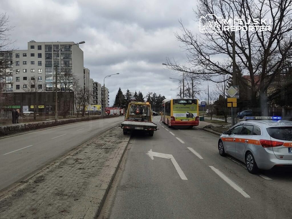 Na drodze stoi laweta z uniesionym pojazdem, obok zaparkowany radiowóz oraz autobus, w tle nowoczesne budynki.