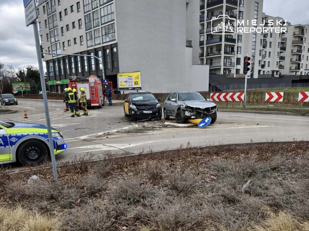 Dwa uszkodzone samochody po kolizji na rondzie, obok stoją strażacy i radiowóz policyjny.