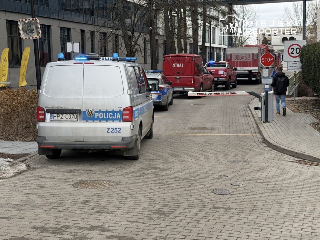 Policyjny furgon z niebieskimi światłami zatrzymany przy wjeździe na teren budynku, obok wozu strażackiego.