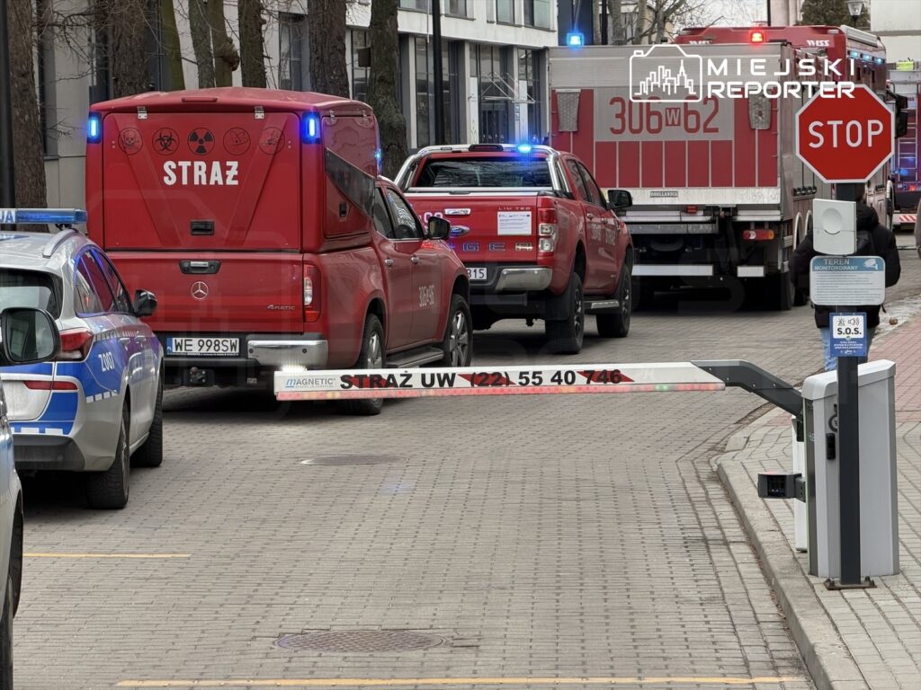 Czerwony wóz strażacki oraz radiowóz policyjny zaparkowane na ulicy, obok znaku "STOP" i szlabanu.