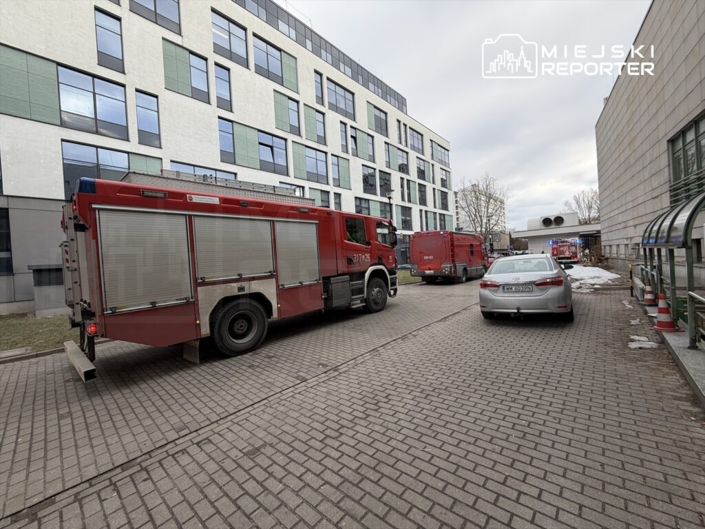 Czerwony wóz strażacki zaparkowany na podjeździe przy nowoczesnym budynku z przeszkloną fasadą.