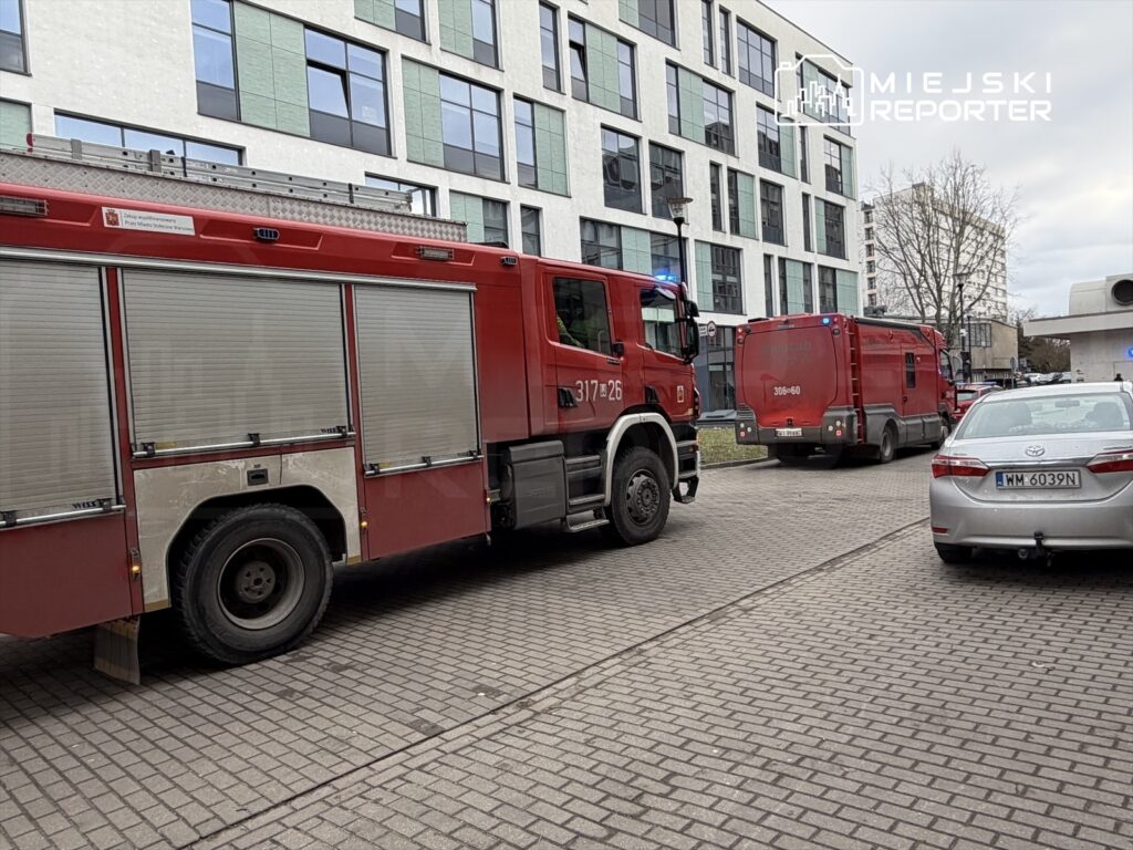 Czerwony wóz strażacki z włączonymi sygnałami świetlnymi parkuje obok innego pojazdu ratunkowego na brukowanej ulicy.