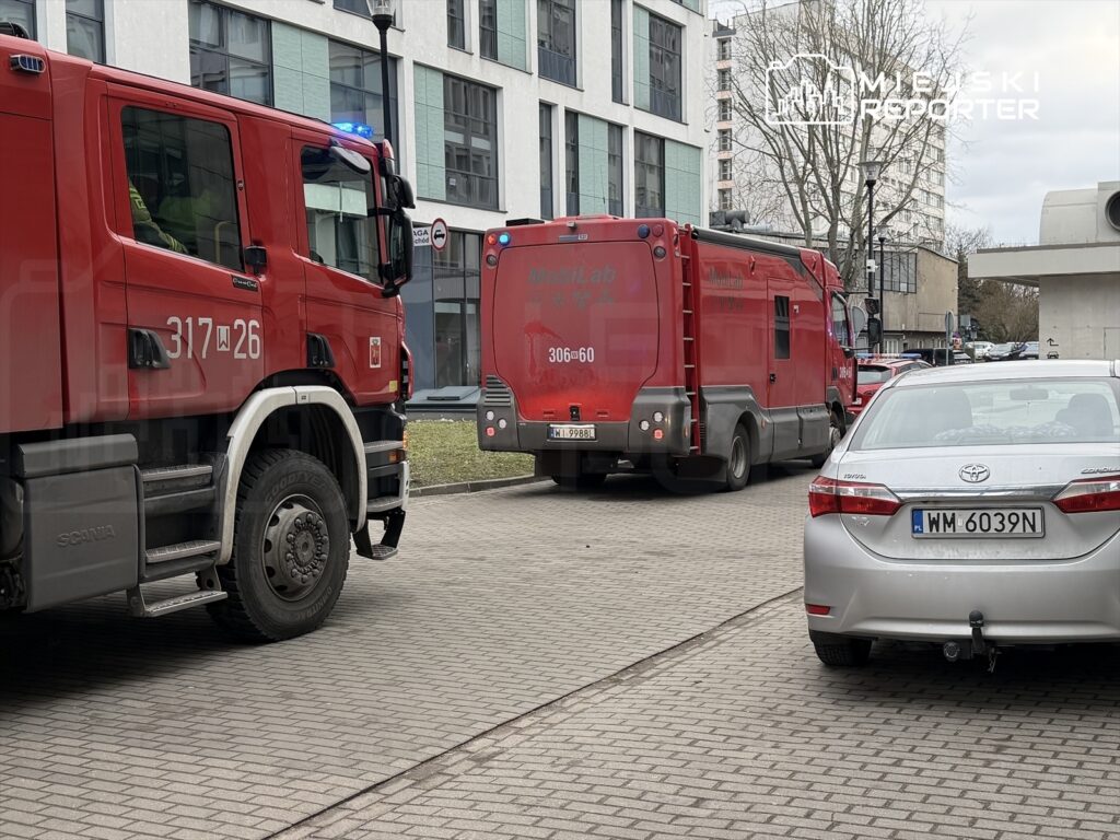 Czerwony wóz strażacki z włączonymi sygnałami świetlnymi stoi na chodniku obok drugiego pojazdu ratunkowego.