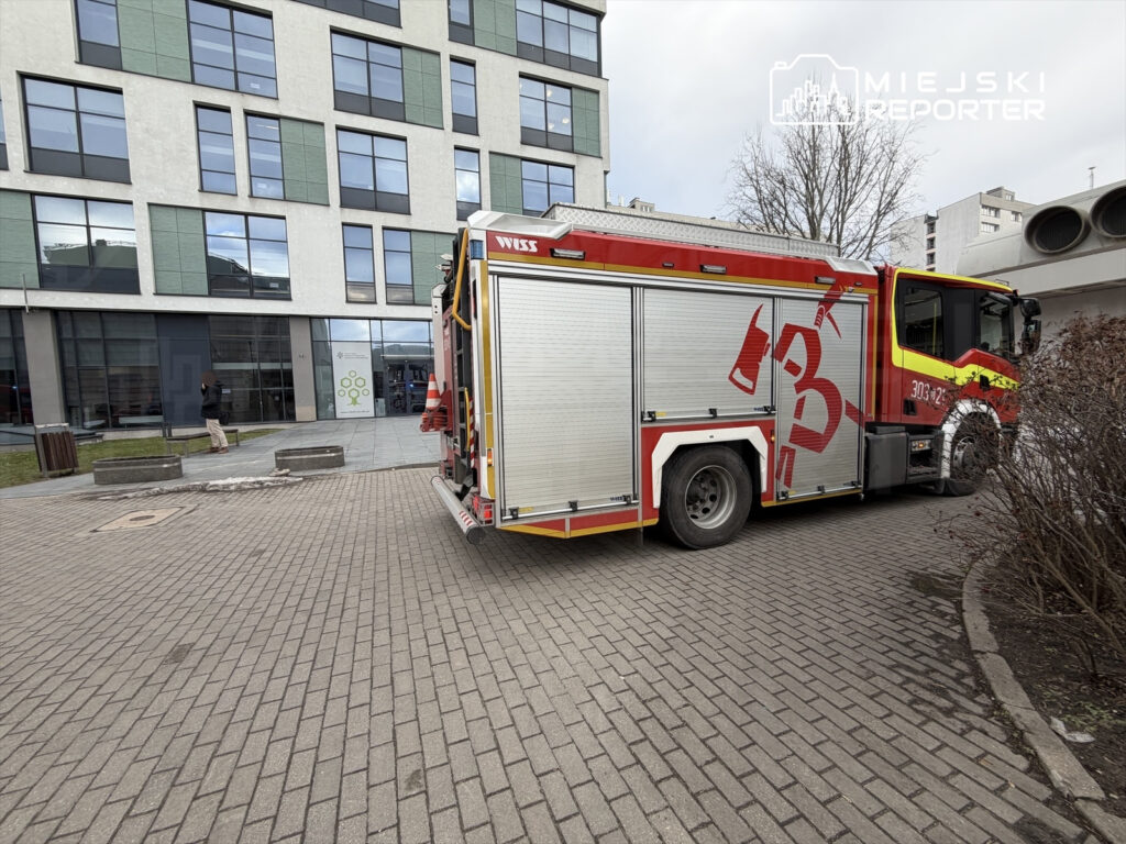 Wóz strażacki zaparkowany na kostkowanej nawierzchni, obok nowoczesnego biurowca z przeszkloną fasadą.