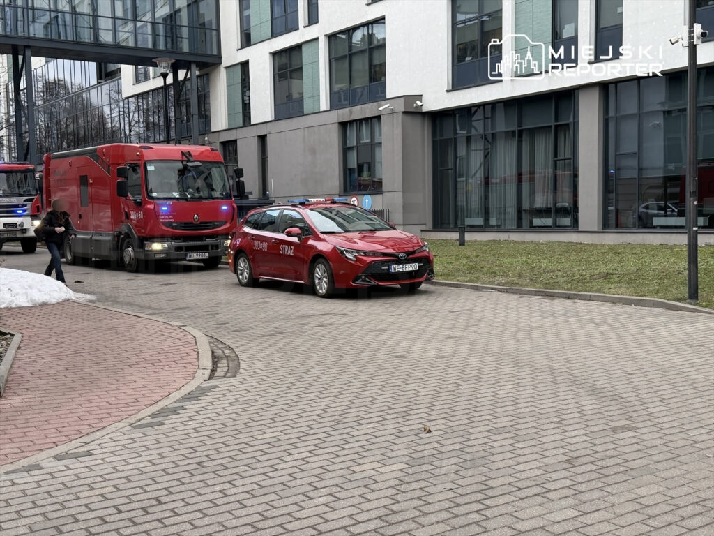 Czerwony samochód strażacki oraz ciężarówka ratunkowa zatrzymane na placu przed nowoczesnym biurowcem.