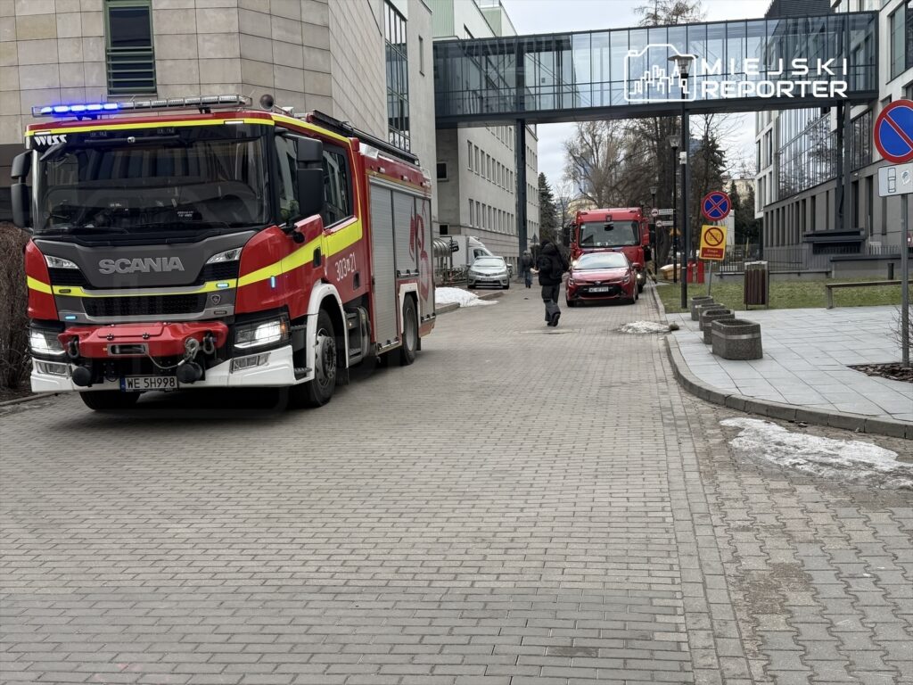 Czerwony wóz strażacki z niebieskimi światłami zatrzymany na chodniku, obok przechodzący mężczyzna w czarnej kurtce.