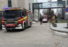 Czerwony wóz strażacki z niebieskimi światłami zatrzymany na chodniku, obok przechodzący mężczyzna w czarnej kurtce.