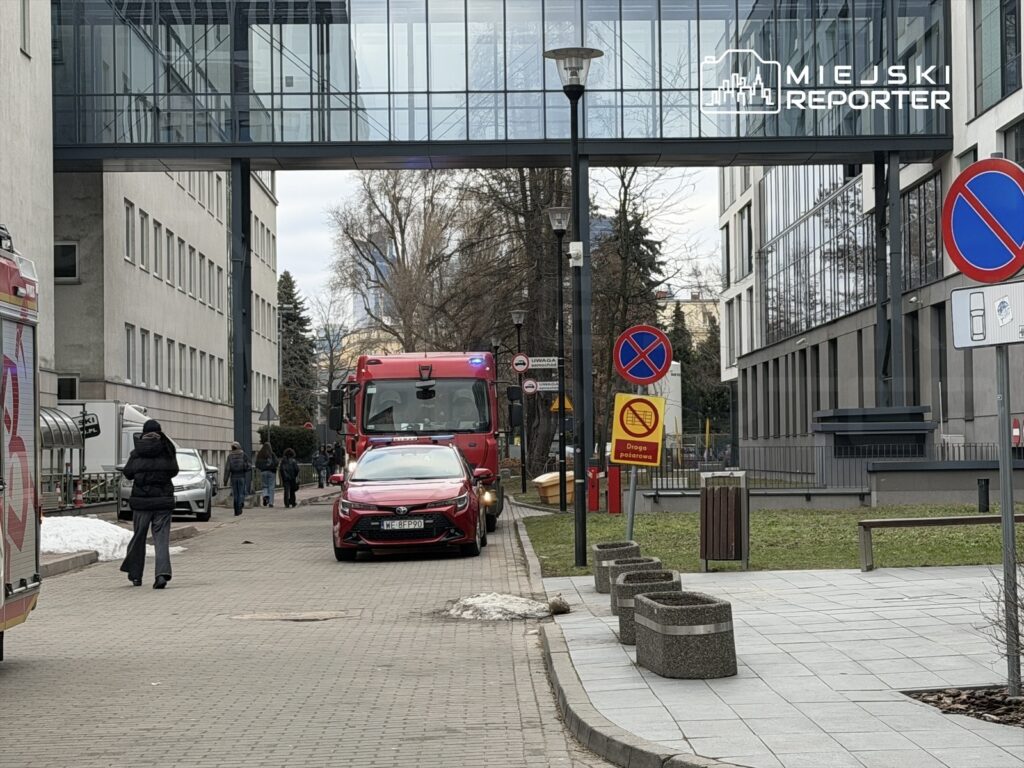 Czerwony wóz strażacki zatrzymany przy nowoczesnym biurowcu, obok przechodnie spacerują chodnikiem.