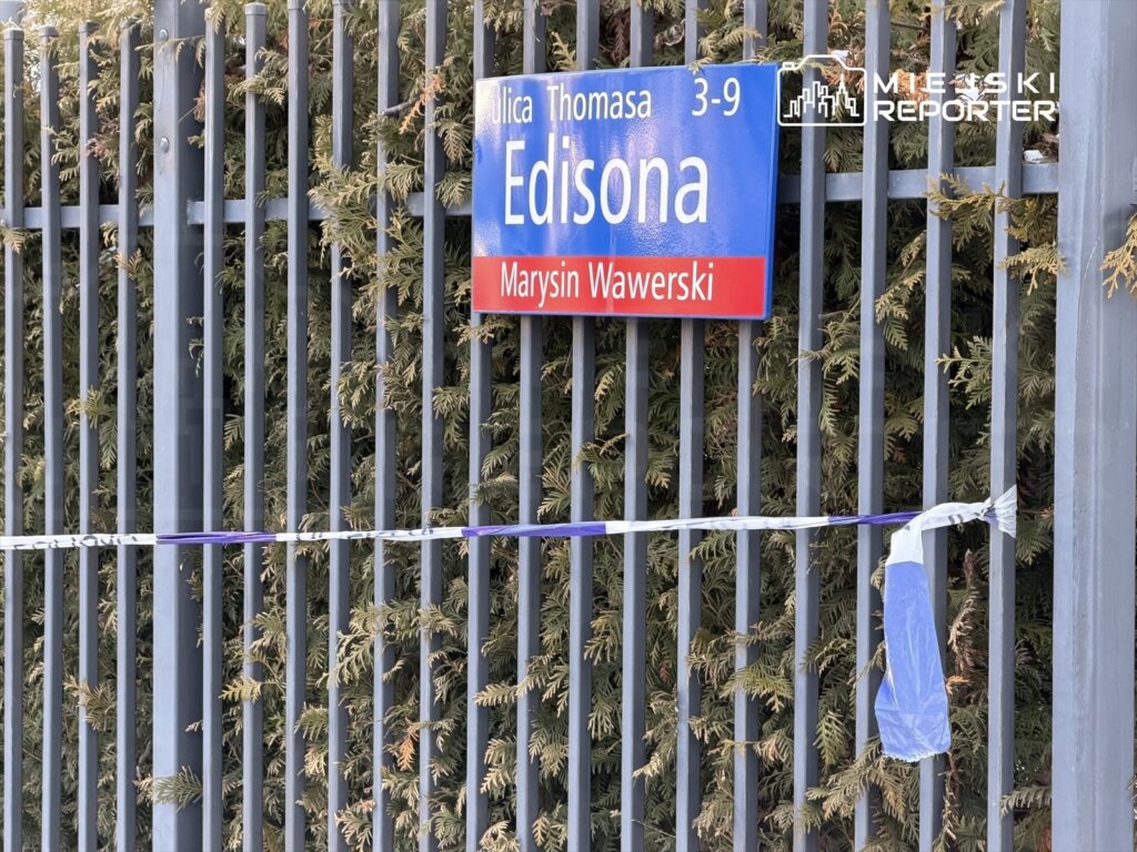Tablica informacyjna z nazwą ulicy "Edisona" oraz numerami 3-9, otoczona zielonymi krzewami i taśmą zabezpieczającą.
