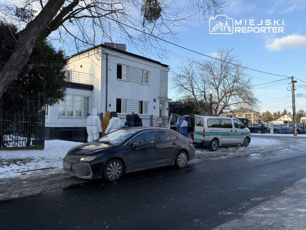 Grupa osób w odzieży ochronnej zbliża się do nowoczesnego, białego budynku, obok parkują policyjny van.