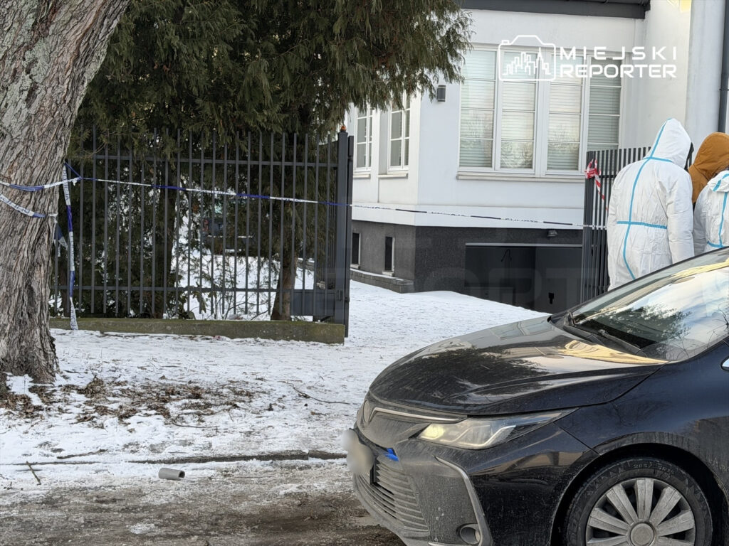 Na zaśnieżonym chodniku stoi czarny samochód, obok grupka ludzi w białych kombinezonach ochronnych, przy ogrodzeniu.