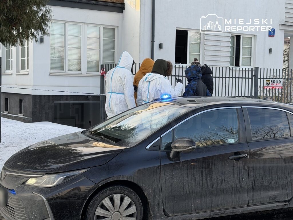 Grupa osób w białych kombinezonach ochronnych rozmawia przy ogrodzeniu przed domem, obok policyjnego samochodu.