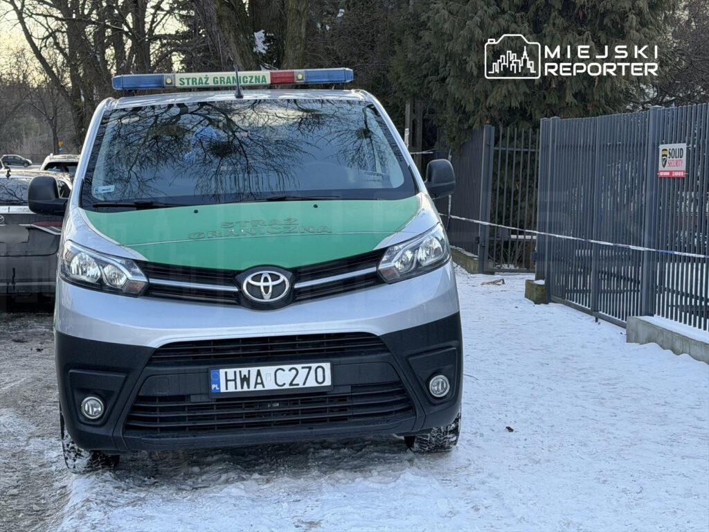 Samochód służbowy Straży Granicznej zaparkowany na zaśnieżonej nawierzchni, w tle ogrodzenie i drzewa.