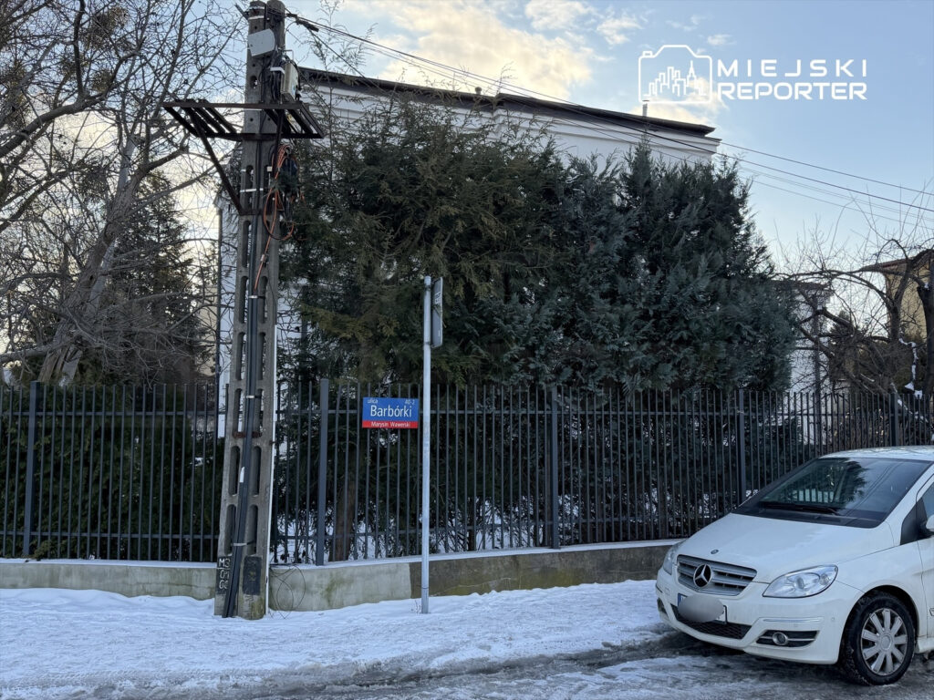 Zimowy krajobraz z białym samochodem zaparkowanym przy ogrodzeniu, obok słupa energetycznego i znaku "Barbórki".