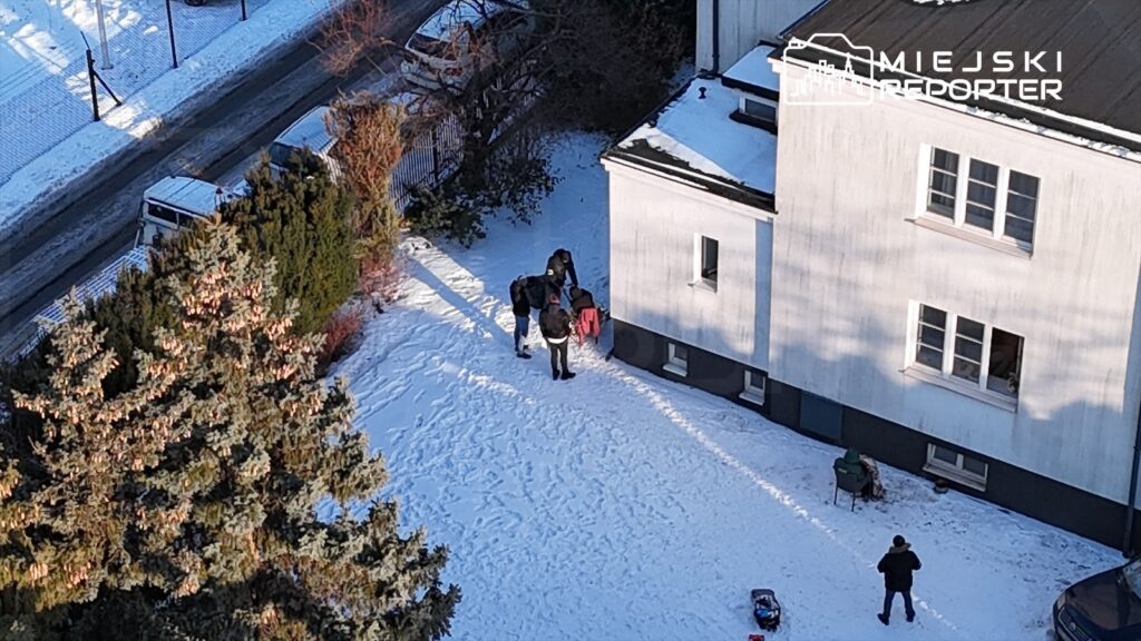 Cztery osoby na zaśnieżonym podwórku rozmawiają i pomagają sobie, obok stoi wózek dziecięcy.