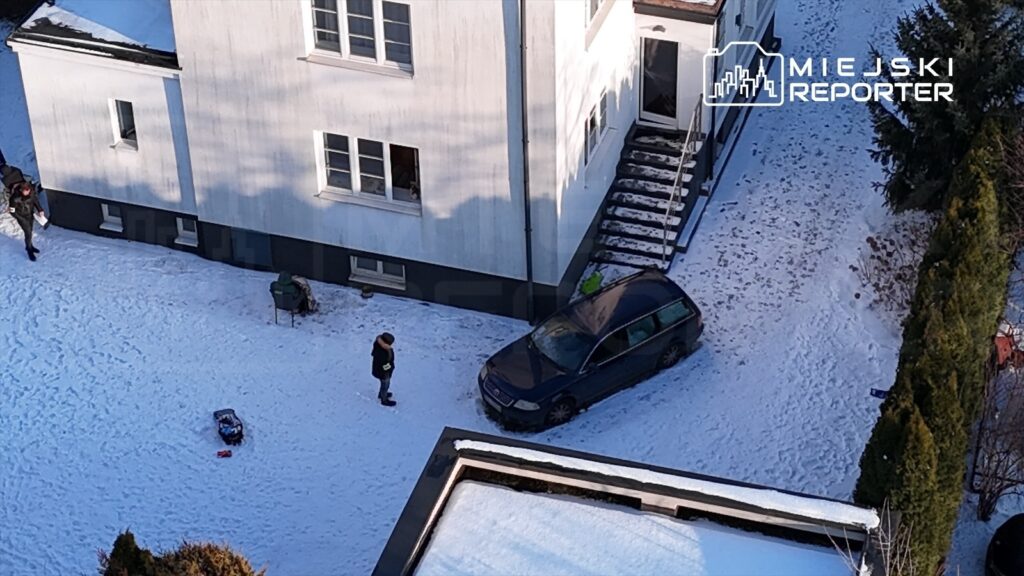 Mężczyzna w czarnej kurtce stoi obok zaparkowanego samochodu na ośnieżonym podjeździe przed budynkiem.