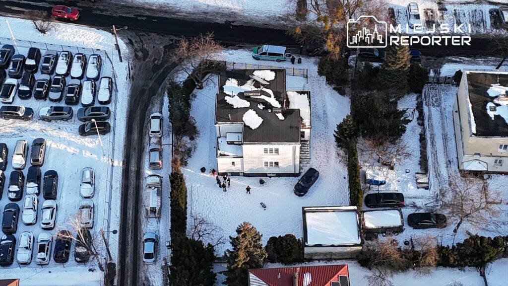Grupa ludzi w zimowej scenerii, bawiąca się na śniegu przed białym domem z płaskim dachem. W tle widoczne samochody na parkingu.