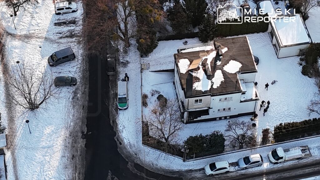 Grupa osób zbiera się na podwórku obok nowoczesnego domu otoczonego śniegiem, podczas gdy samochody zaparkowane są w pobliżu.